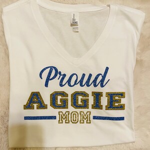 May include: White t-shirt with a blue and gold glitter design that says "Proud Aggie Mom".