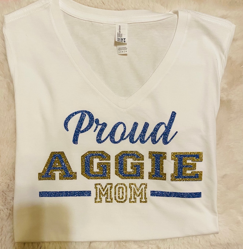 May include: White t-shirt with a blue and gold glitter design that says "Proud Aggie Mom".