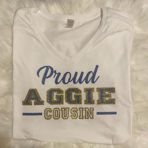 May include: White V-neck t-shirt with the text "Proud AGGIE COUSIN" in blue and gold glitter lettering. The shirt is folded and displayed on a white, fluffy surface. The text is centered on the shirt.