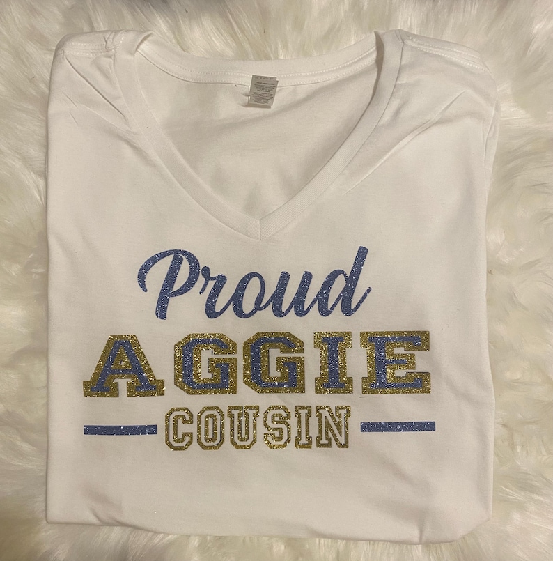 May include: White V-neck t-shirt with the text "Proud AGGIE COUSIN" in blue and gold glitter lettering. The shirt is folded and displayed on a white, fluffy surface. The text is centered on the shirt.