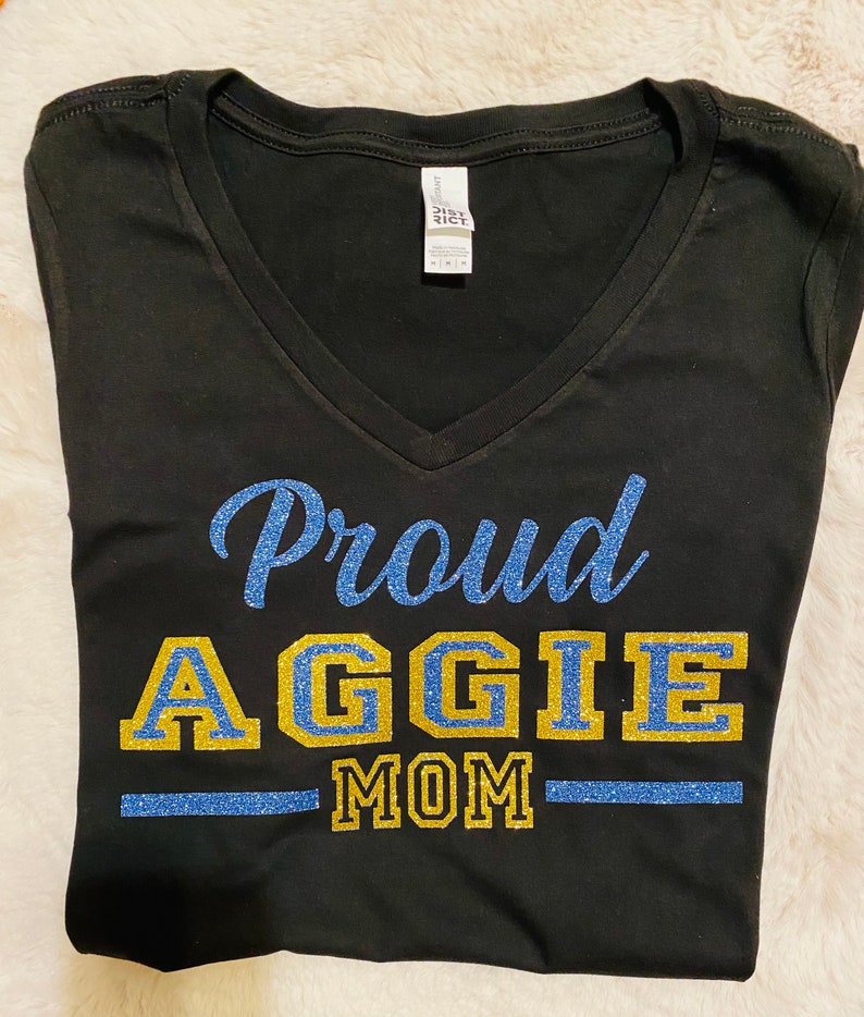May include: Black v-neck t-shirt with a blue and gold glitter design that says "Proud Aggie Mom".
