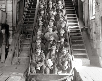 May include: A black and white historical photograph showing a large group of miners wearing hats with lamps, seated in rows inside a wooden mine transport car. The men are positioned on an incline, with one individual standing to the left side.
