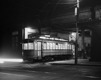 Peut inclure: Une scène nocturne en noir et blanc montrant un tramway d'époque éclairé de l'intérieur, stationné près d'une structure. Le tramway affiche le texte GILMOR ST à l'avant. Un lampadaire lumineux éclaire les rails au-dessus.