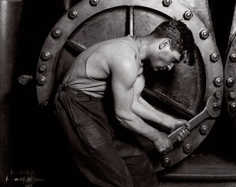 Könnte beinhalten: Ein Schwarz-Weiß-Foto, das eine Person in einem Unterhemd und einer Hose zeigt, die mit einem großen Metallschlüssel eine Schraube an einer massiven Industriemaschine festzieht. Die Maschine hat ein großes kreisförmiges Gehäuse mit Bolzen.