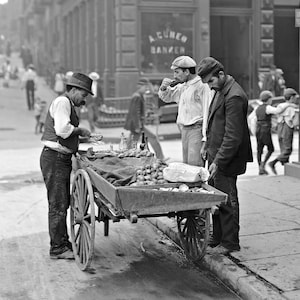 May include: A black and white photo of a street vendor selling produce from a cart. The vendor is wearing a vest and a hat. Two men are standing near the cart. One man is drinking from a cup. The sign on the building behind the vendor reads 