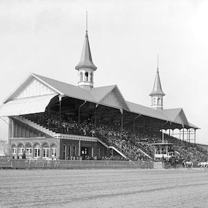 May include: A black and white photograph of a large, multi-level grandstand at a horse racing track. The structure features two prominent spires, arched windows on the lower level, and a sloped roof covering a crowd of spectators in the stands.