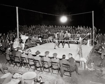 May include: A black and white photograph of a boxing match taking place in an outdoor ring at night. Two boxers are sparring in the center of the ring while a referee stands between them. A large crowd of spectators watches from the surrounding area.
