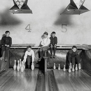 May include: A black and white photograph of several young people acting as pinsetters in a bowling alley. They are positioned behind wooden bowling lanes, manually setting up white bowling pins. The wall behind them features large painted numbers.