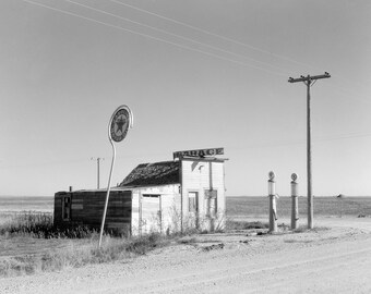 May include: A black and white photograph of a weathered wooden garage building with a sign reading GARAGE on the roof. A tall Texaco Gasoline Motor Oil sign stands to the left, and two vintage fuel pumps stand near a wooden utility pole.