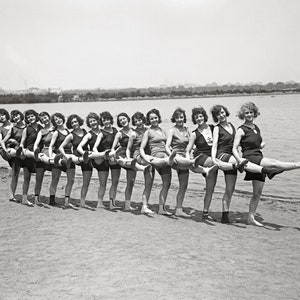 Könnte beinhalten: Eine Gruppe von Frauen in Vintage-Badebekleidung steht an einem Sandstrand, jede mit einem ausgestreckten Bein in einer synchronisierten Pose. Sie lächeln alle und schauen in die Kamera. Die Frauen tragen einteilige Badeanzüge in verschiedenen Stilen und Mustern. Der Strand ist sandig und im Hintergrund befindet sich Wasser.
