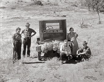 May include: A black and white photograph of a vintage automobile with the text OREGON OR BUST painted on the rear. The vehicle is heavily loaded with bundled gear and supplies, parked in a dry, grassy field with four individuals standing or sitting nearby.