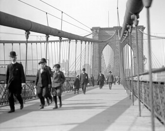 May include: A black and white photograph showing the Brooklyn Bridge pedestrian walkway with several individuals walking across. The scene features the bridge's iconic stone towers, suspension cables, and metal railings in a historic setting.
