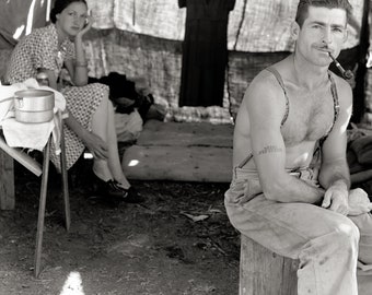 May include: A black and white photograph showing a person sitting on a wooden block wearing trousers with suspenders and smoking a pipe, while another person sits in the background wearing a checkered dress and dark shoes near a metal pot.