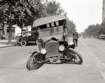 May include: A vintage black automobile with a soft top and a damaged front axle, causing the front wheels to splay outward. The vehicle features a prominent radiator grille, round headlights, and a license plate reading 28 104 DC.
