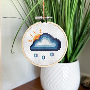 Puede incluir: Un aro de bordado blanco con un diseño de punto de cruz de una nube azul con un sol amarillo asomando por detrás. Gotas de lluvia caen de la nube.