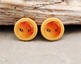 May include: Two circular, light tan pendants featuring a central orange, sun-like circle with a black, almond-shaped eye graphic. Each pendant has a small metal loop at the top for attachment, showcasing a smooth, flat, and round design.