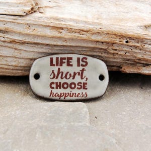 May include: A white ceramic oval tag with the text "LIFE IS short, CHOOSE happiness" in red lettering. The tag has two holes on either side.