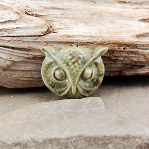 May include: A green ceramic owl pendant with brown speckles and large eyes. The owl is hanging from a silver loop.