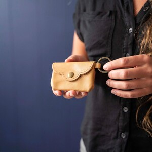May include: A small, light tan leather pouch with a scalloped flap and a single brass-colored snap button closure. The pouch features a circular metal key ring attached to the side, providing a functional way to secure keys or attach to bags.