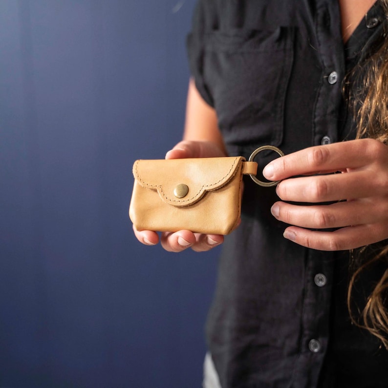 May include: A small, light tan leather pouch with a scalloped flap and a single brass-colored snap button closure. The pouch features a circular metal key ring attached to the side, providing a functional way to secure keys or attach to bags.