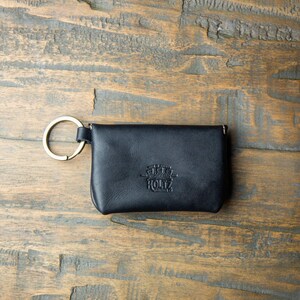 May include: A black leather pouch with a gold-toned metal key ring attached to the side. The front of the pouch features a debossed logo that reads Made in America Holtz Leather Co. The pouch has a rectangular shape with rounded corners.