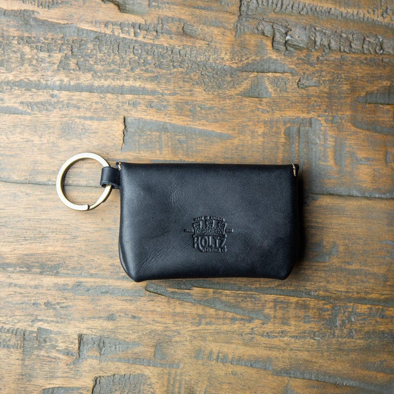 May include: A black leather pouch with a gold-toned metal key ring attached to the side. The front of the pouch features a debossed logo that reads Made in America Holtz Leather Co. The pouch has a rectangular shape with rounded corners.