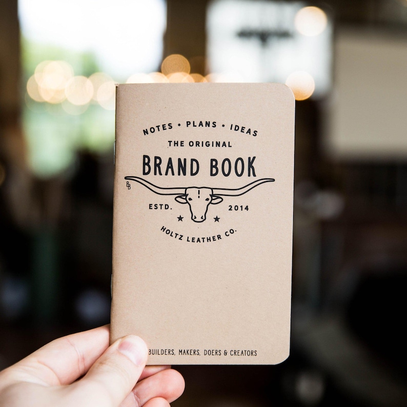 May include: A tan notebook with black text and a longhorn steer graphic. The cover reads: NOTES PLANS IDEAS THE ORIGINAL BRAND BOOK ESTD. 2014 HOLTZ LEATHER CO. BUILDERS, MAKERS, DOERS & CREATORS. The notebook has a simple, rectangular paper cover.