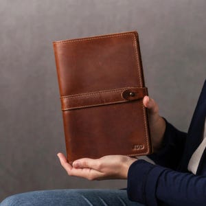 May include: A rectangular brown leather notebook cover featuring a horizontal strap with a single metal snap button closure. The front surface displays the embossed letters JDD in the bottom right corner, showcasing a smooth, matte texture.