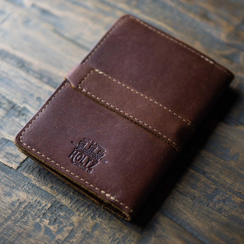 May include: A dark brown leather passport holder featuring a horizontal strap across the front and visible light-colored stitching. The cover is embossed with the text HOLTZ LEATHER CO and MADE IN AMERICA, showcasing a smooth, matte finish.