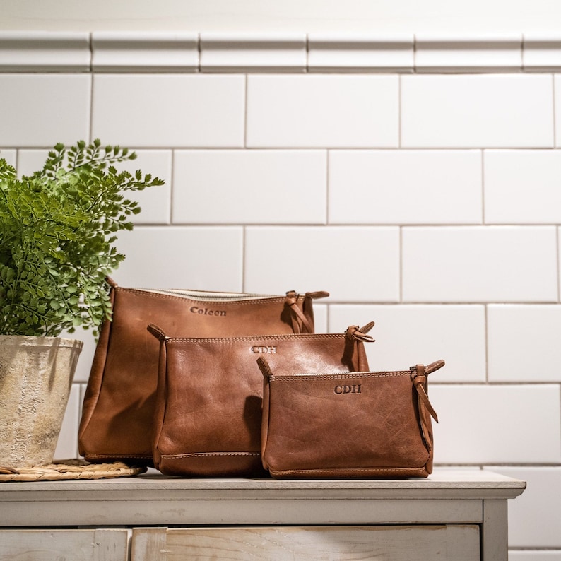 May include: Three brown leather pouches of varying sizes arranged in a row. The largest pouch is embossed with the name Coleen, while the two smaller pouches are each embossed with the letters CDH. Each pouch features a zipper closure and leather pulls.