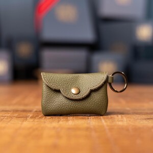 May include: A small olive green leather pouch with a pebbled texture, featuring a scalloped flap closure secured by a single brass-colored metal snap button. A circular metal key ring is attached to the right side of the rectangular accessory.