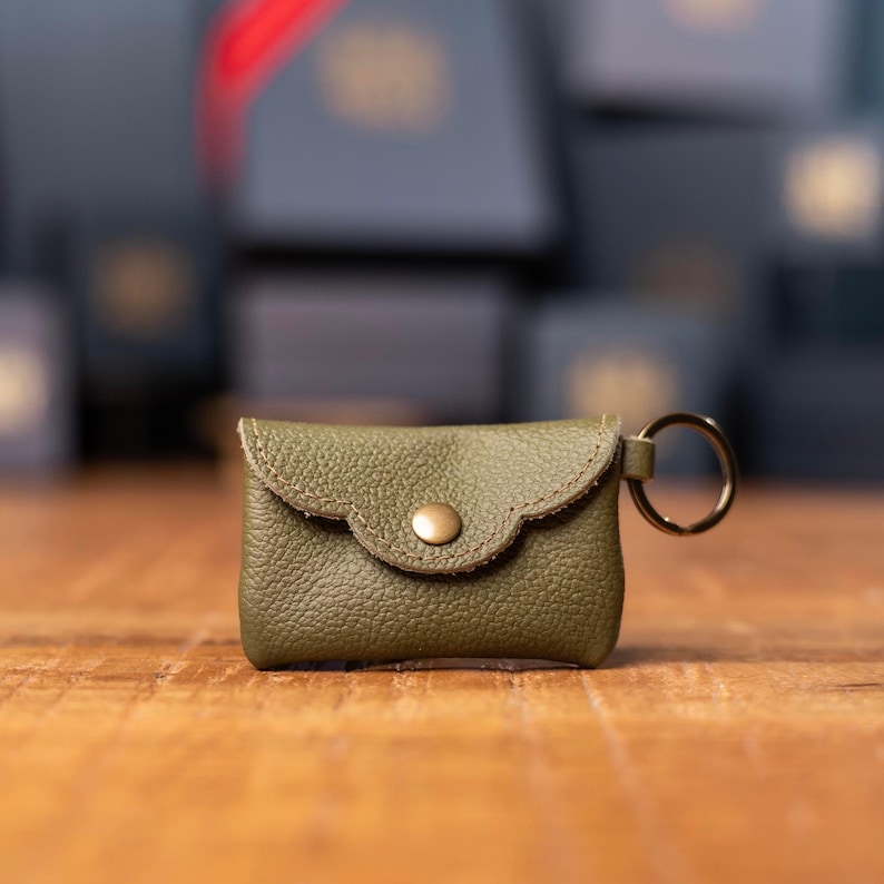 May include: A small olive green leather pouch with a pebbled texture, featuring a scalloped flap closure secured by a single brass-colored metal snap button. A circular metal key ring is attached to the right side of the rectangular accessory.