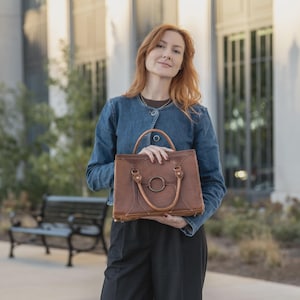 May include: A brown leather handbag with a circular metal accent and top handles. The bag has a structured design and is held by a person wearing a denim jacket and black pants. The background features a building and greenery.