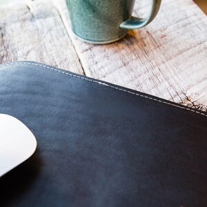 May include: A smooth, dark black leather desk mat featuring rounded corners and a clean, light-colored stitched border along the edge. The surface has a matte finish and is positioned flat, partially covered by a white computer mouse.