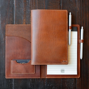 May include: A tan leather notebook cover with a smooth finish, featuring a vertical pocket, a business card slot, and two pen loops. The cover is embossed with the letters SAJ in the bottom right corner and holds a lined notepad with a Holtz logo.
