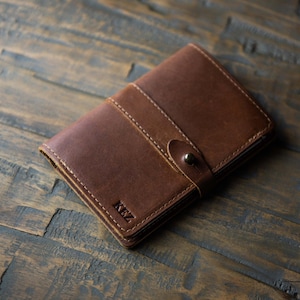 May include: A brown leather notebook cover featuring a vertical strap closure with a metal snap button. The cover has visible stitching along the edges and a strap, with the letters KEZ embossed in the bottom corner of the front panel.