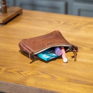 May include: A brown leather zippered pouch with the letters CDH embossed on the front. The pouch is partially open, revealing a pack of Extra gum, a purple lip balm tube, and an orange-capped marker or pen stored inside the main compartment.
