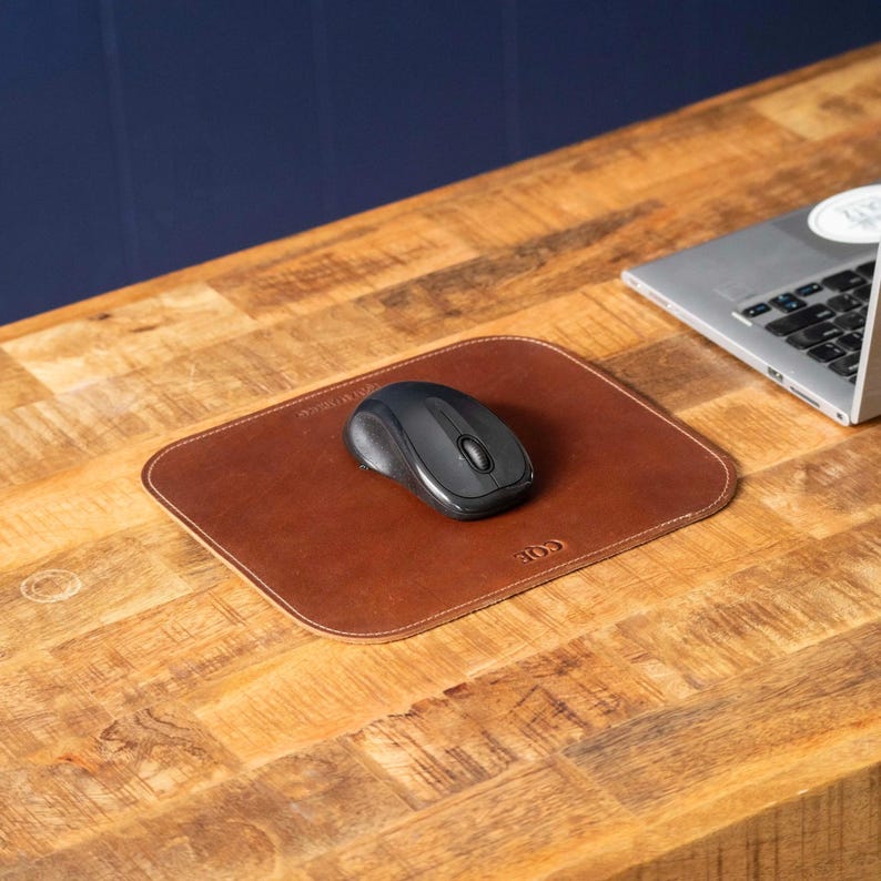 May include: A rectangular brown leather mouse pad with rounded corners and a debossed logo, featuring a black wireless computer mouse centered on its surface. The mouse pad has visible stitching along the edges and a smooth, matte finish.