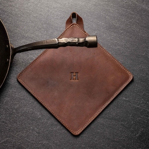 May include: A square brown leather pot holder featuring a centered embossed letter H and a small leather hanging loop at the top. A dark metal pan handle with a textured, hammered finish extends horizontally across the top of the leather square.