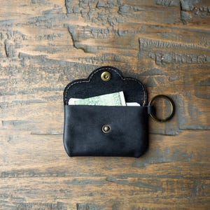 May include: A black leather pouch with a rounded flap, secured by a brass-colored snap button. The pouch features a matching brass-colored metal key ring attached to the side. Inside the open pouch, a small piece of paper currency is visible.