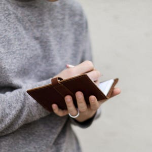 May include: A brown leather notebook with a secure strap closure, held open to reveal white pages. The notebook features visible stitching along the edges and a smooth, matte finish, providing a compact surface for writing with a pencil.