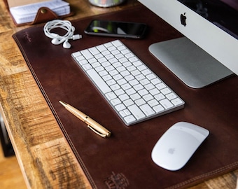May include: A dark brown leather desk mat with stitched edges and a debossed logo in the corner. The mat holds a white wireless keyboard, a white wireless mouse, a gold-toned ballpoint pen, a smartphone, white wired earbuds, and a small notebook.