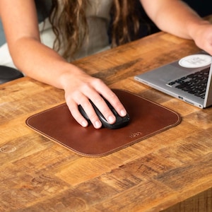 May include: A square brown leather mouse pad with rounded corners and a small embossed logo in the bottom right corner. The mouse pad features a smooth surface and stitched edges, providing a flat base for a black computer mouse.