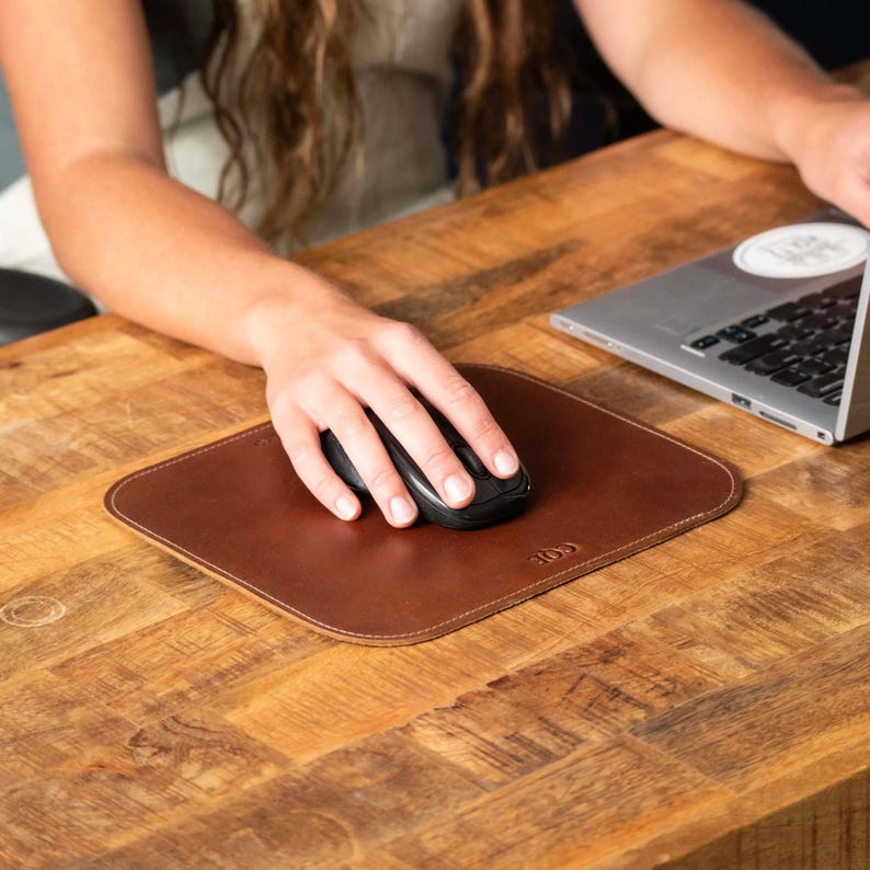 May include: A square brown leather mouse pad with rounded corners and a small embossed logo in the bottom right corner. The mouse pad features a smooth surface and stitched edges, providing a flat base for a black computer mouse.