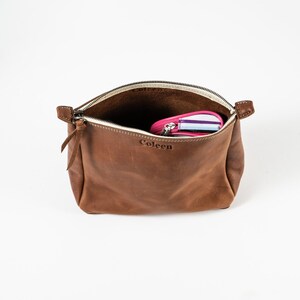 May include: A brown leather pouch with a top zipper closure and a leather pull tab. The name Coleen is embossed on the front. The interior is open and contains a small pink and white striped zippered case visible inside the main compartment.