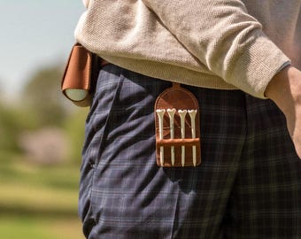 May include: A tan sweater worn over navy blue plaid trousers with a brown leather golf accessory attached to the waistband. The accessory features a pouch for a golf ball and a separate holder containing four white golf tees in a vertical row.