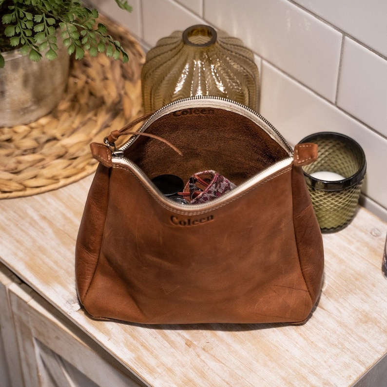 May include: A brown leather pouch with a zipper closure and the name Coleen embossed on the front. The interior is open and contains small items. The pouch has a structured base and a smooth, matte finish, showcasing natural leather textures.