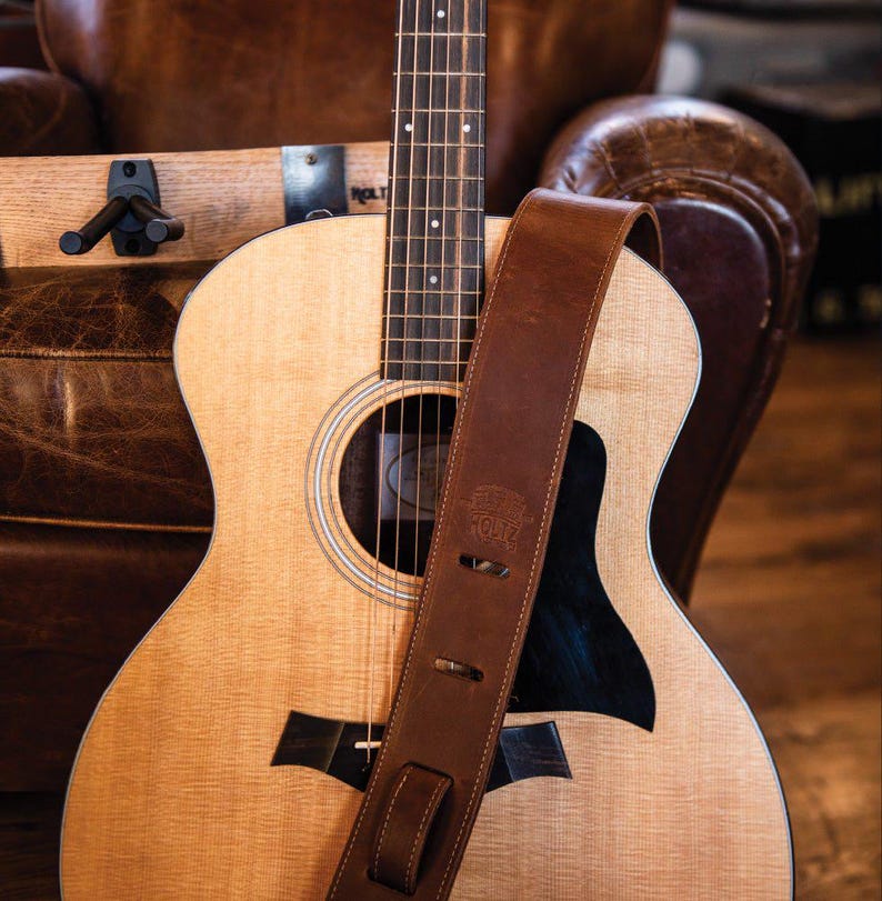 May include: A light brown leather guitar strap with visible stitching and a stamped logo rests across the body of an acoustic guitar. The guitar features a light-colored wood top, a dark pickguard, and a dark fretboard with white dot inlays.