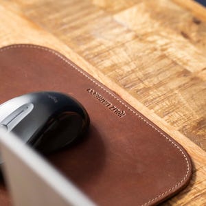 May include: A brown leather mouse pad with rounded corners and white stitching along the edges. The text HOLTZ LEATHER CO is embossed on the surface. A black computer mouse is positioned on the left side of the leather pad.