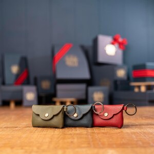 May include: Three small envelope-style pouches made of textured leather, each featuring a gold-tone snap button closure and a metal keyring attached to the side. The pouches are colored olive green, dark gray, and bright red, arranged in a row.